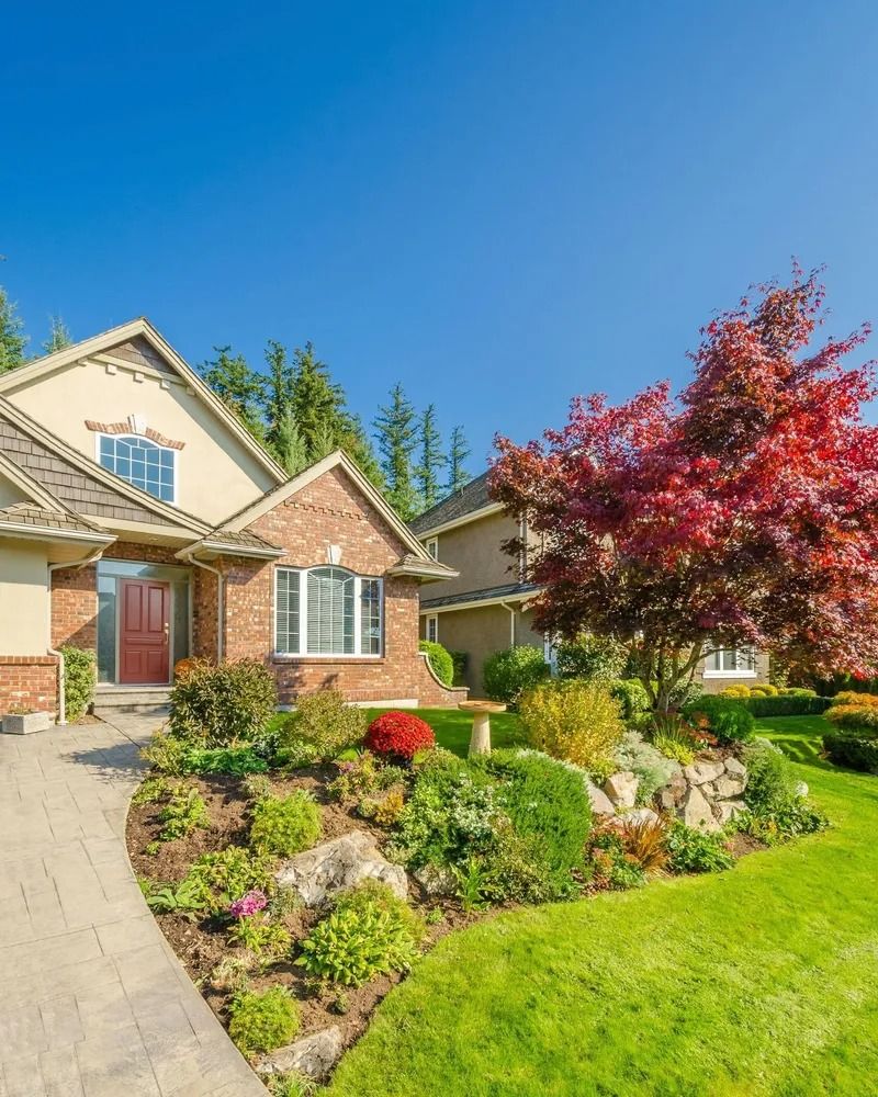Traditional suburban house with brick accents and an elegant front yard featuring a bright red maple tree, a curved walkway, and a beautiful rock garden landscape.