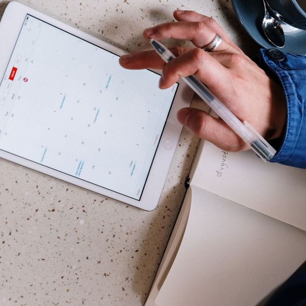 A person's hand, wearing a ring, holds a pen and points to a digital calendar displayed on a tablet, showing the month of August. An open notebook with "August" written on it is next to the tablet. A blue cup and saucer are partially visible in the upper r