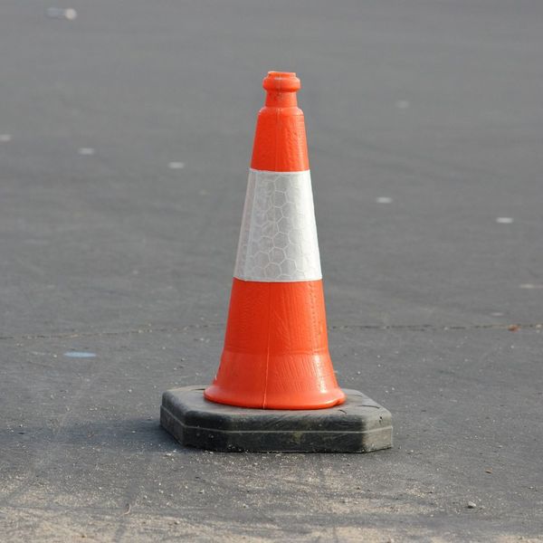 A bright orange traffic cone with a white reflective stripe sits on a black square base on an asphalt surface.