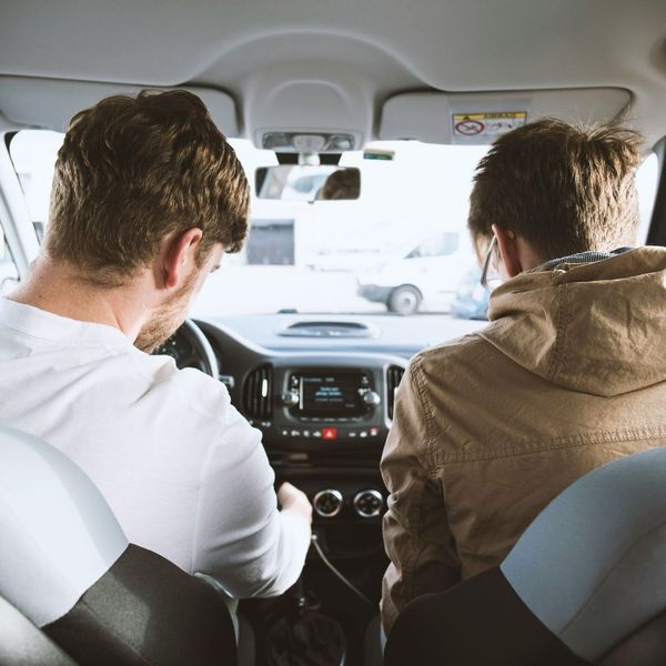 A view from the back seat of a car showing two individuals in the front seats. The person in the driver's seat (left, facing away) has brown hair and is wearing a white t-shirt. The person in the passenger seat (right, facing away) has brown hair and is we