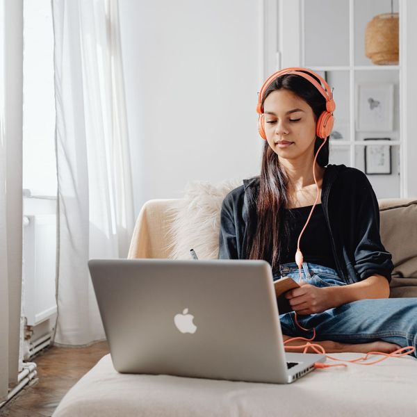 A young woman with long dark hair, wearing a black long-sleeved shirt and jeans, sits comfortably on a couch. She is wearing orange headphones and looking at a silver laptop in front of her while holding a small notebook and pen.