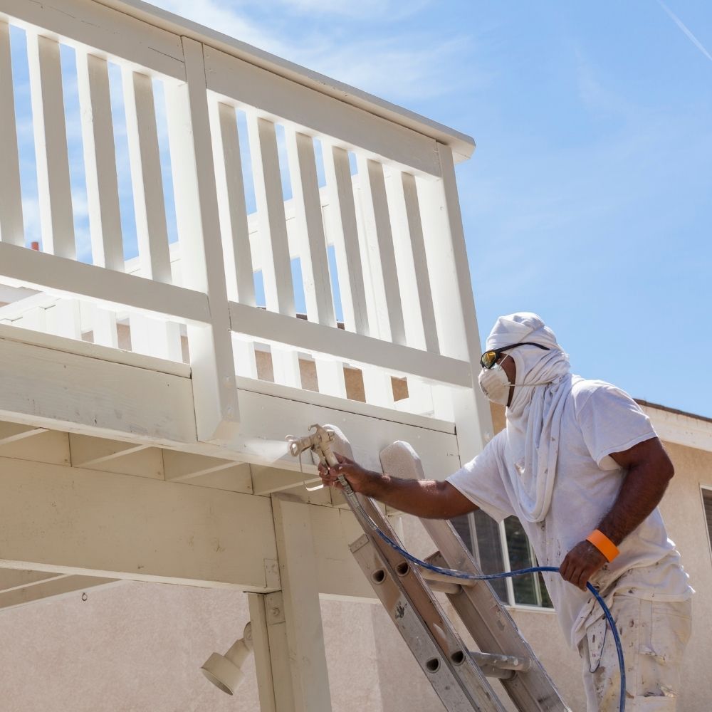 Painter in protective gear on a ladder using a paint sprayer to apply white paint to an exterior deck.