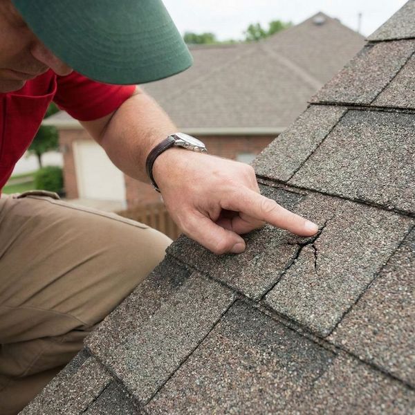 A close-up photograph of a professional roofing inspector pointing to a small crack in a shingle on a residential roof.