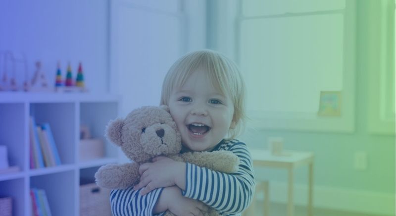 A happy toddler hugging a small teddy bear in a bright, inviting room.