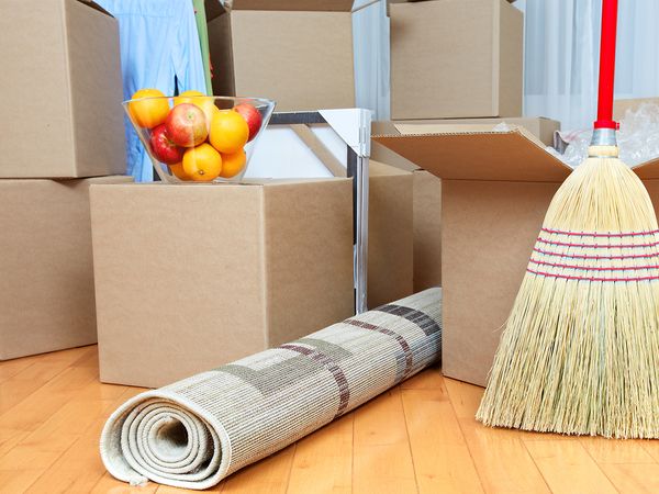 Several moving boxes stacked in a room with a rug, a broom, and a bowl of fruit