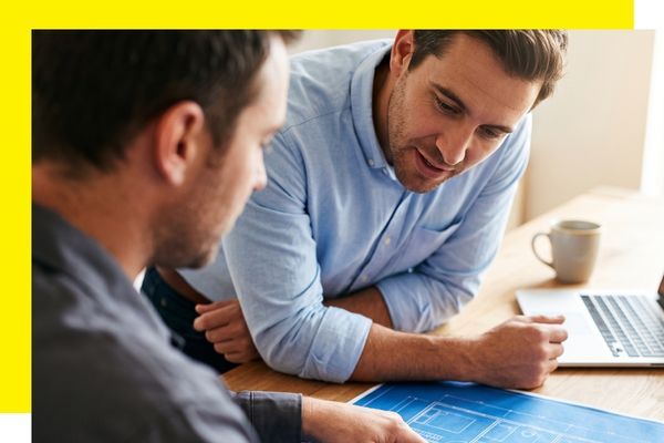 Two men in collared shirts leaning over a wooden desk to review blue building blueprints.