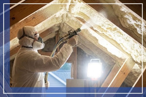 A professional applicator in a full protective suit and respirator actively spraying expanding foam insulation between wooden rafters in a residential attic space.