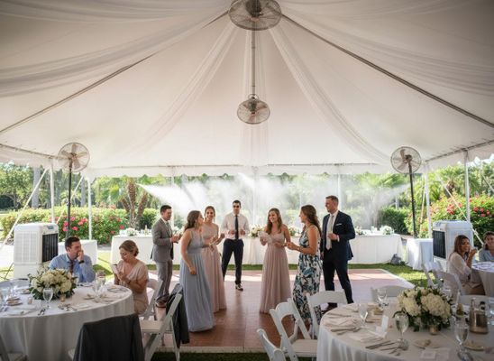 Guests mingle and chat at an outdoor wedding reception under a large white tent with cooling misters and fans. Outdoor Wedding Reception Under Tent with Guests and Misters