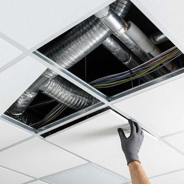 a section of a suspended drop ceiling with several acoustic tiles removed, showing neatly arranged HVAC ducts, electrical wiring, and plumbing conduits accessible within the dark plenum space above a modern commercial building