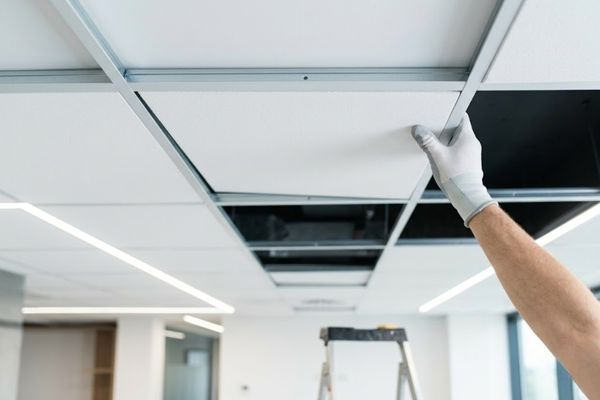a professional's hand precision-fitting a crisp white acoustic tile into a T-bar main runner grid during a suspended drop ceiling installation in a clean, modern office space