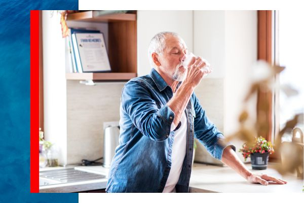 An older man leaning against a countertop and drinking a glass of water