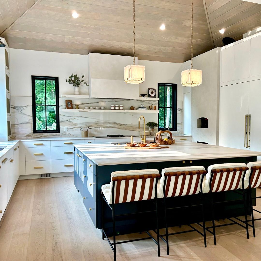 Luxury kitchen with a wood vaulted ceiling, custom white cabinetry, dark oversized island, marble countertops and backsplash, brass fixtures, and upholstered counter stools. Luxury Contemporary Kitchen with Vaulted Ceiling