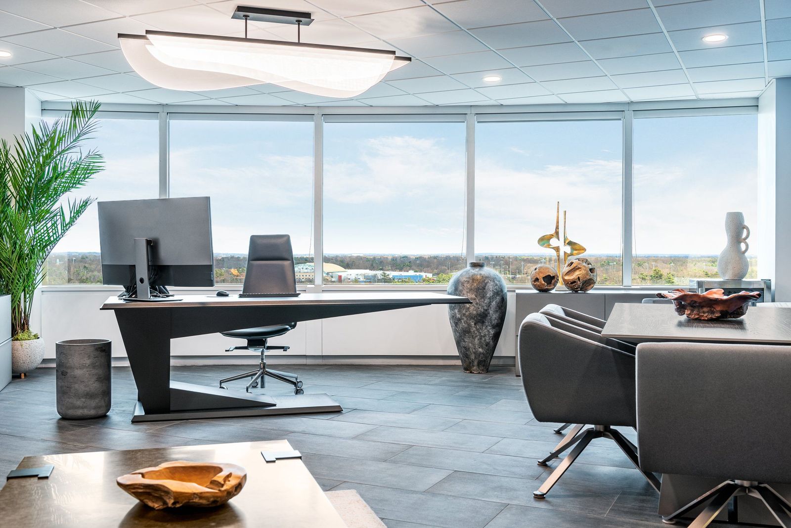 Executive office designed by Serenella Interiors, featuring a custom-fabricated statement desk inspired by an iconic television drama, floor-to-ceiling windows with panoramic views, a sculptural pendant light, slate flooring, and curated sculptural art objects that give the space a commanding, editorial presence. Custom Executive Desk Design in a High-Rise CEO Office | Serenella Interiors