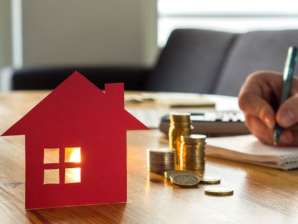 A red paper cutout of a house, a stack of coins, and someone writing in a notebook. 