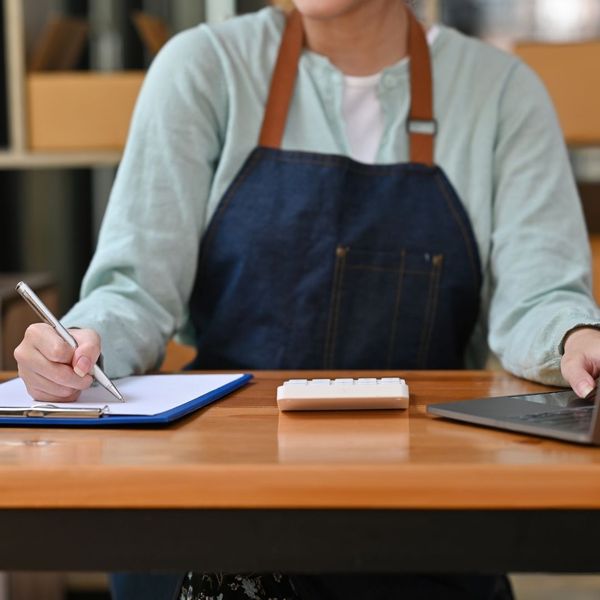 worker in overalls writing on clipboard, using laptop with other hand, calculator on desk