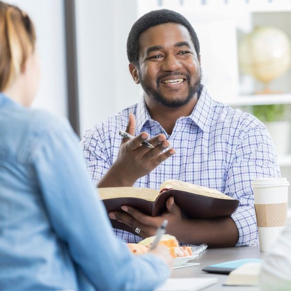 Man reading from a Bible and gesturing to someone across the table. 