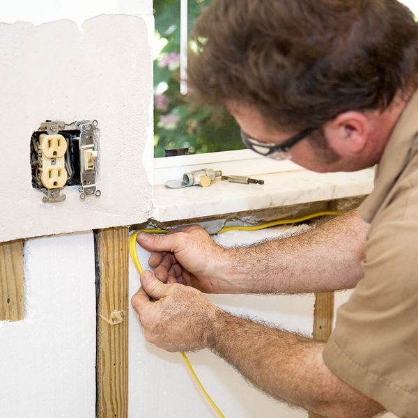 A construction worker in a hard hat and safety glasses inspects exposed bathroom pipes and wiring with a flashlight during a remodel. remodel.jpg