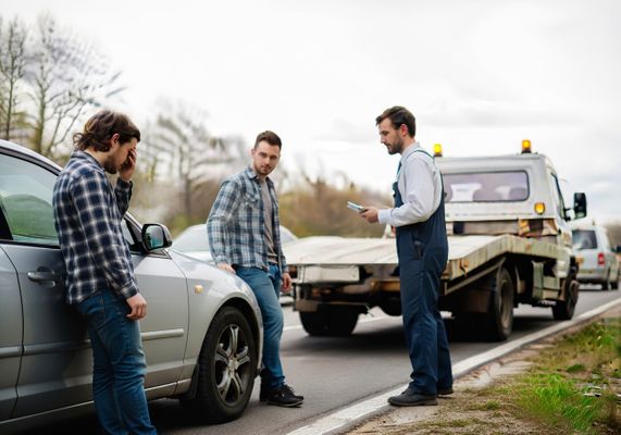 Two men stand beside a damaged silver car, one with his hand on his head in distress, while a tow truck driver with a clipboard stands nearby assessing the situation on a roadside. Aftermath of a Car Accident