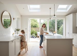 A woman chops vegetables in a bright kitchen with a child drawing at the counter, featuring skylights and garden access. Woman and child in a modern kitchen with garden view