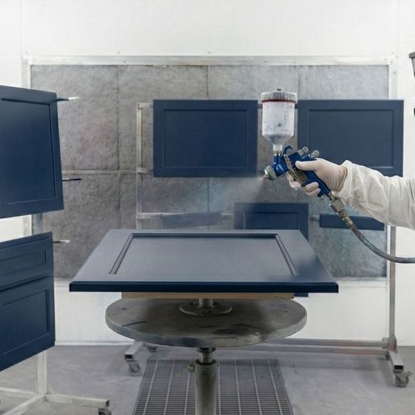 A professional painter in a white protective suit using a spray gun to apply a smooth navy blue finish to cabinet doors in a shop environment. A professional painter in a white protective suit using a spray gun to apply a smooth navy blue finish to cabinet doors in a shop environment.
