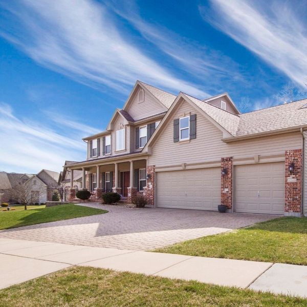 A large, two-story suburban house with light beige siding, brick accents, and a wide driveway, seen on a bright day with wispy clouds.