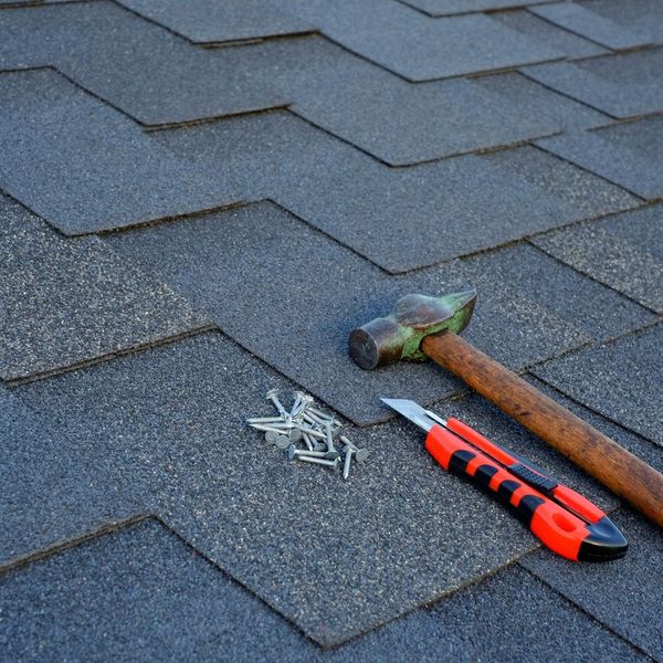 A close-up of a dark gray asphalt shingle roof with roofing tools including a hammer, a utility knife, and a pile of roofing nails placed on the shingles.