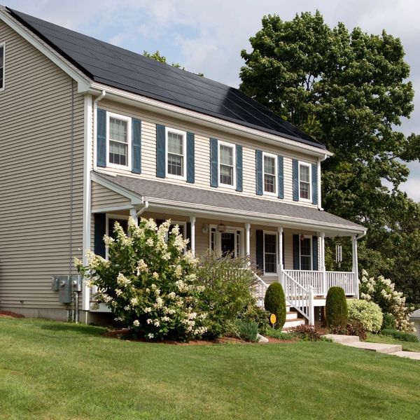 A two-story traditional American house with beige siding, blue shutters, a front porch, and solar panels installed on its dark roof, surrounded by green lawn and trees.
