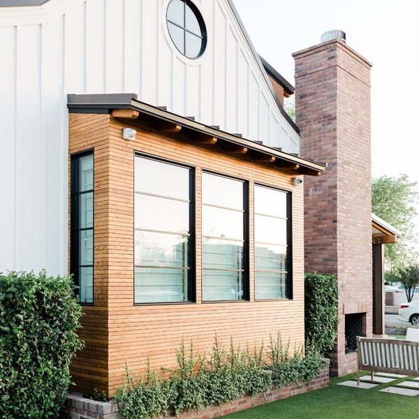 The modern exterior of a house featuring white vertical siding, a round window, natural wood paneling around large windows, and a brick chimney with outdoor seating.