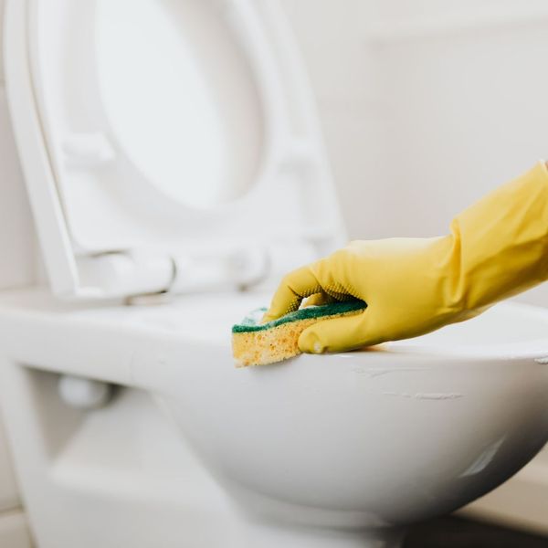 A gloved hand (yellow glove) using a sponge to clean the base of a white toilet bowl.