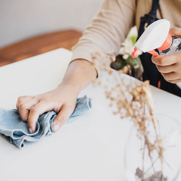 A person's hand wiping a white tabletop with a blue cloth, while holding a spray bottle in the other hand. A vase with dried flowers is partially visible. A person's hand wiping a white tabletop with a blue cloth, while holding a spray bottle in the other hand. A vase with dried flowers is partially visible.