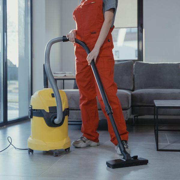 A person in orange overalls using a large yellow wet/dry vacuum cleaner on a tiled floor in a modern living room. A person in orange overalls using a large yellow wet/dry vacuum cleaner on a tiled floor in a modern living room.