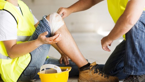 Construction worker with bandaged knee sitting on floor, another construction worker inspecting the bandage