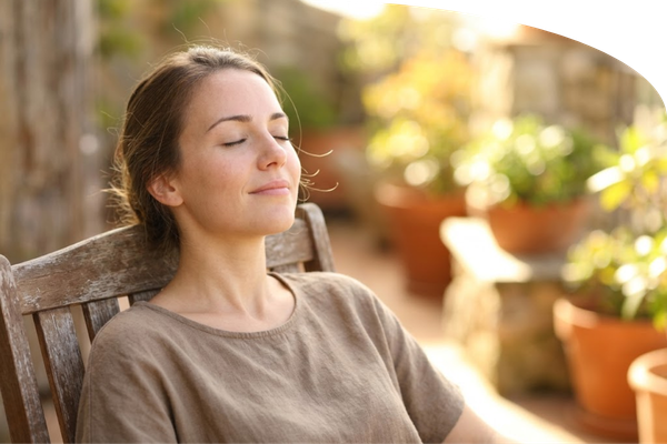 a young woman sitting peacefully on a sun-drenched patio, eyes closed and smiling softly while taking a deep breath of fresh air