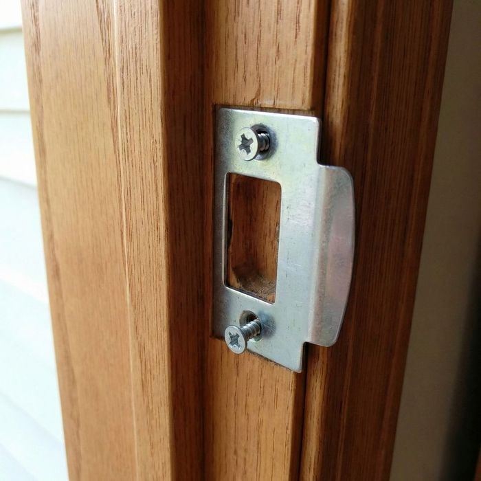 A close-up photograph of a metal strike plate on a wooden door frame where the mounting screws are loose and not flush with the surface.