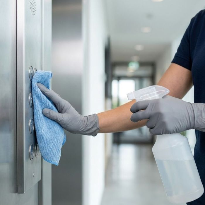A close-up photograph of a gloved hand wiping down a stainless steel elevator button panel with disinfectant.