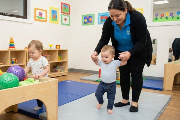 A teacher in the Rainforest Child Care Center young toddlers classroom helps a child learn to walk, while another toddler plays at a sensory table. A teacher in the Rainforest Child Care Center young toddlers classroom helps a child learn to walk, while another toddler plays at a sensory table.