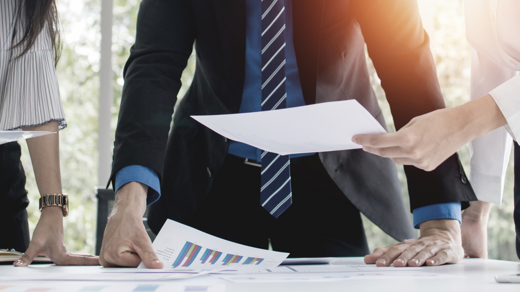 Three formally dressed people look over documents in a bright conference room