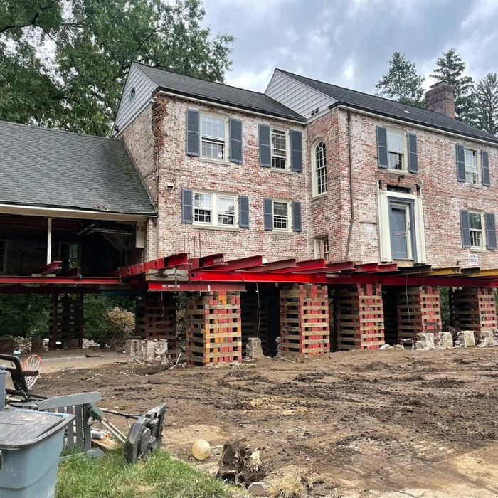 A large brick house is elevated on red steel beams and extensive wooden cribbing on a construction site.