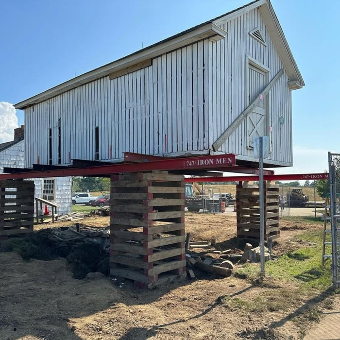 A tall, white, barn-style building is elevated high on red steel beams and extensive wooden cribbing on a construction site.