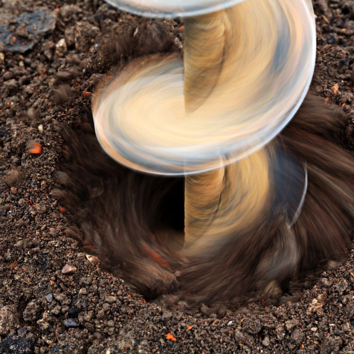 A large metal auger with helical flights drills into dark soil, creating a hole for a foundation pile.