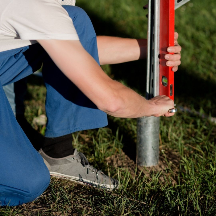 A construction worker in blue overalls kneels, using a bright orange spirit level to ensure a metal foundation post is perfectly vertical in the ground.
