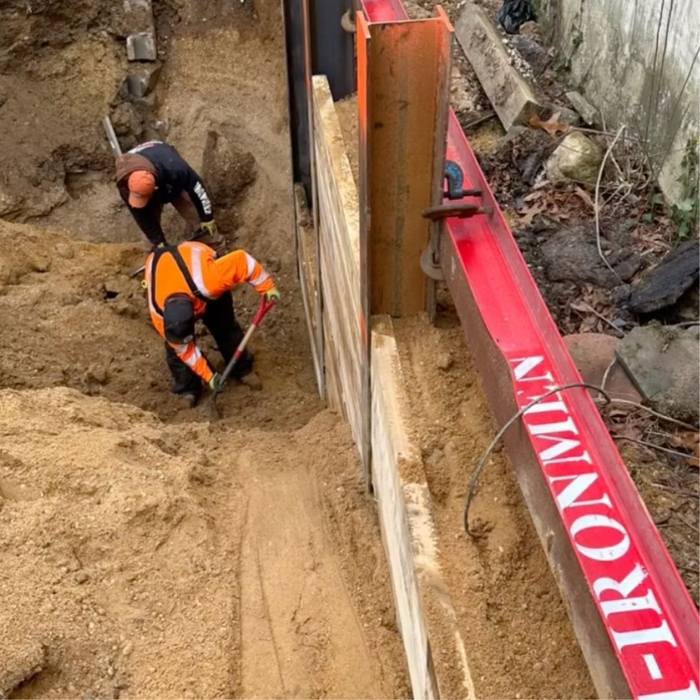 Two construction workers in safety gear are working in a narrow trench next to a long red metal beam with "IRONMEN" visible on it.