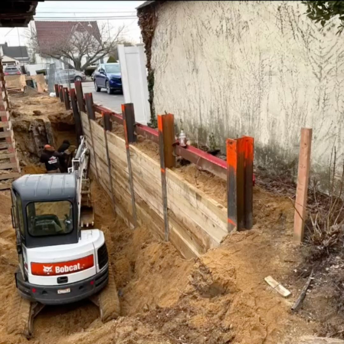A small white excavator is positioned next to a deep trench reinforced with wooden shoring, on a construction site.