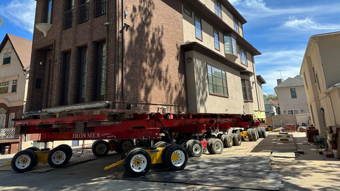 A large brick house is elevated on red steel beams and extensive wooden cribbing on a construction site.