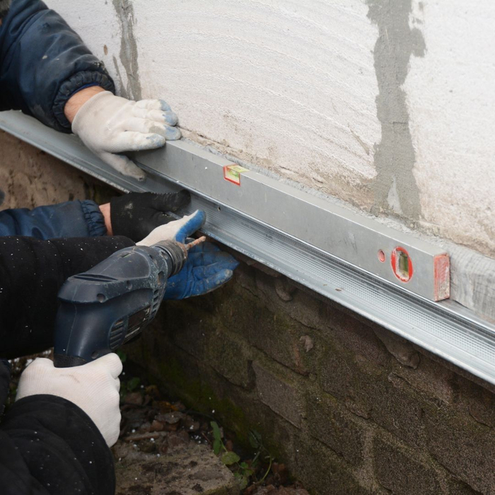 A worker's gloved hands use a level against a metal beam being installed along a foundation wall.