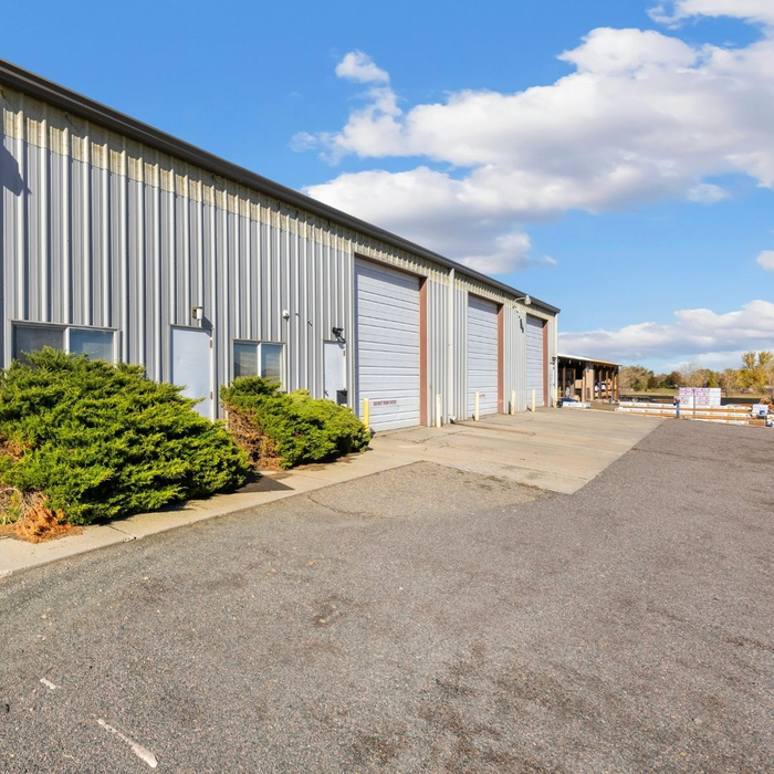 A row of grey metal commercial buildings with roll-up doors and a paved driveway under a partly cloudy sky.