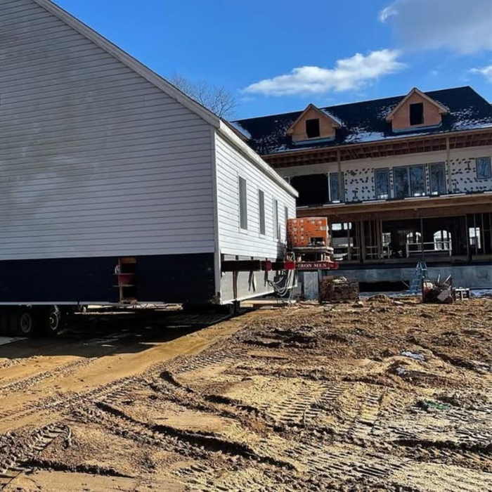 A light-colored house is being carefully moved along a dirt path on specialized equipment next to a new construction site.