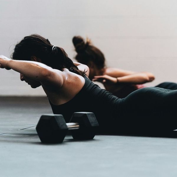 Two women performing back extension exercises (supermans) on yoga mats during a corporate fitness class in a bright, industrial-style studio.