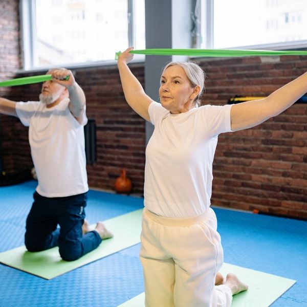 An active senior couple participating in a group fitness class, using green resistance bands for overhead mobility exercises.