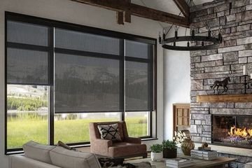 a rustic living room with a stone fireplace and large windows covered by dark, black-out roller shades.